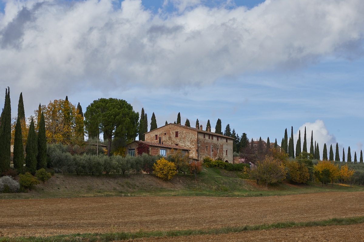 A rustic building is set against a backdrop of rolling hills and a partly cloudy sky. Cypress trees line the scene, complementing the autumn foliage in shades of green, yellow, and orange. A field of cultivated land stretches in the foreground, highlighting the rural landscape.