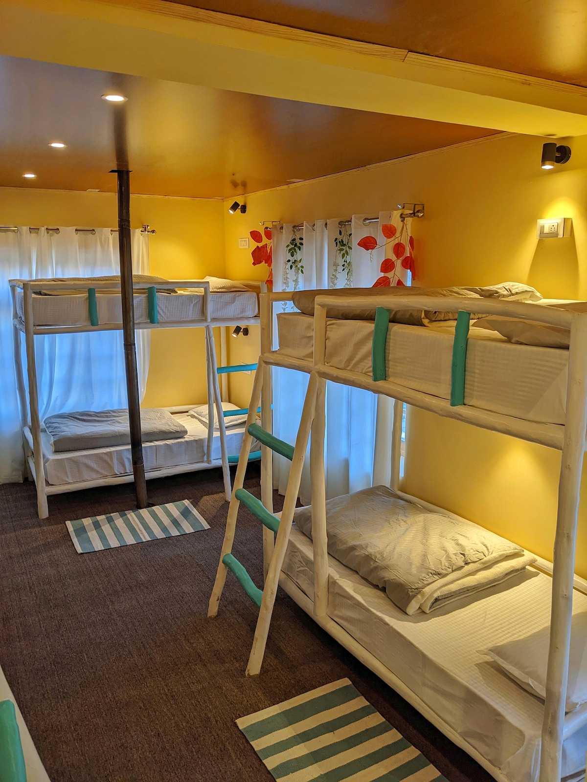 The bunk room features two sets of white wooden bunk beds arranged along the yellow walls, contributing to a bright atmosphere. Light grey bedding is neatly arranged on each mattress. Soft light is provided by ceiling fixtures, highlighting the cozy space, while striped rugs are placed on the floor.
