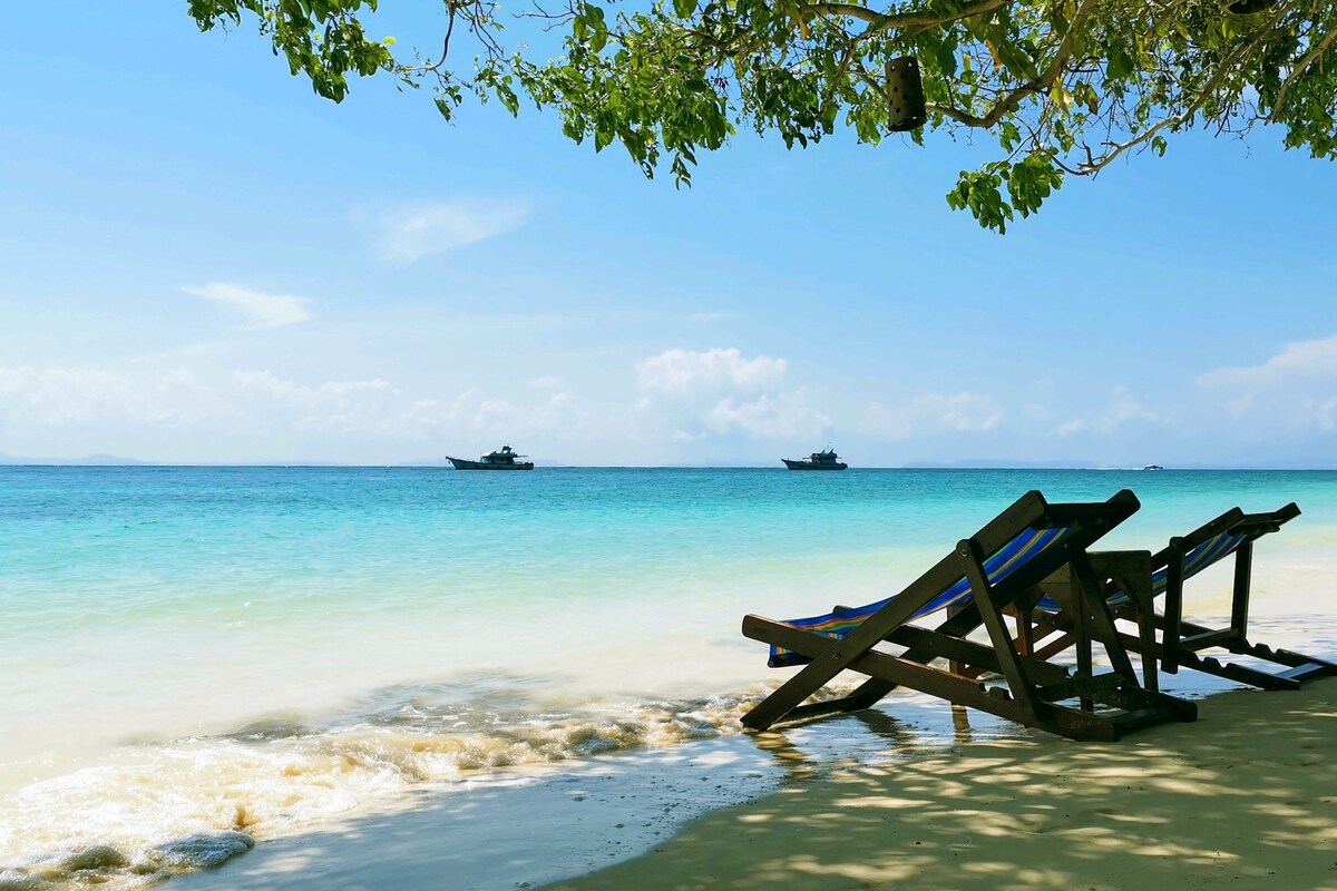 A serene beach scene features wooden lounge chairs positioned on soft, white sand. Gentle waves lapping at the shore create a tranquil atmosphere, while two boats are seen in the distance against a backdrop of clear blue skies and vibrant turquoise water.