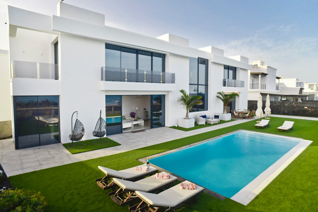 A modern villa showcases a spacious terrace and an inviting swimming pool, surrounded by a well-kept lawn. Two stylish hanging chairs are positioned beside the pool, with sun loungers arranged nearby, providing a relaxed outdoor setting overlooking the sea.