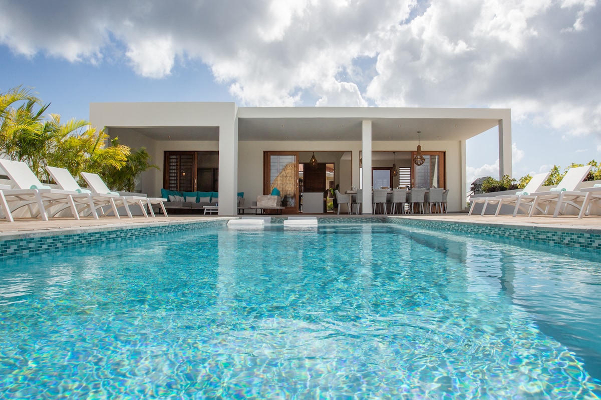 A private pool area is highlighted by lounge chairs surrounding the clear blue water. A spacious outdoor dining area is visible under the covered porch, complemented by views of lush greenery. The villa's modern architecture is framed by clouded skies.