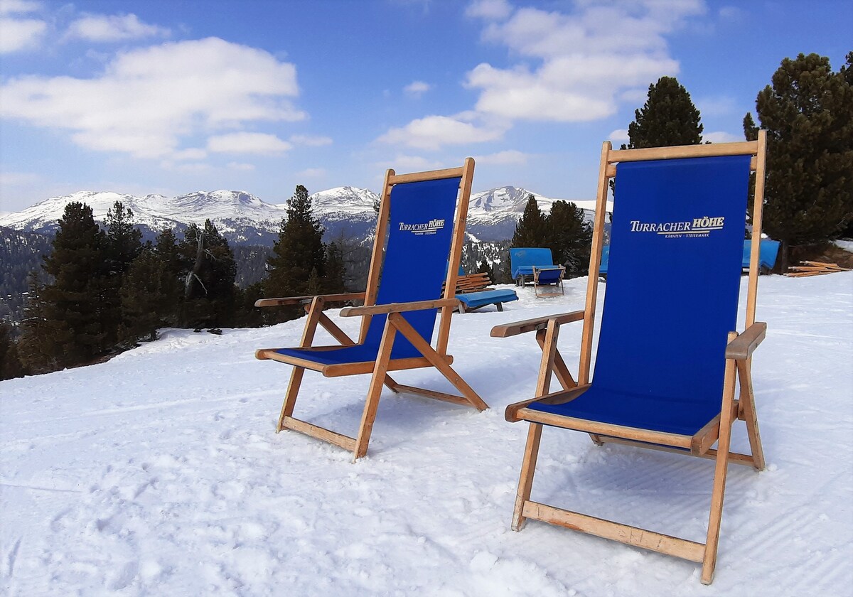 Two blue deck chairs with wooden frames are positioned on a snowy landscape. The scenic backdrop features mountains covered in snow under a partly cloudy sky, offering a serene winter setting.