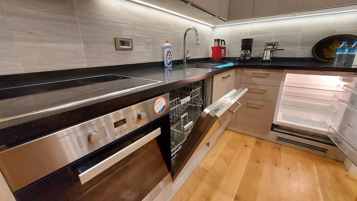 A modern kitchen features sleek cabinetry and a black granite countertop. Stainless steel appliances include a dishwasher and an oven, while a refrigerator is positioned to the right. A bottle of cleaning solution and a blue cloth are placed near the sink, complementing the overall design.