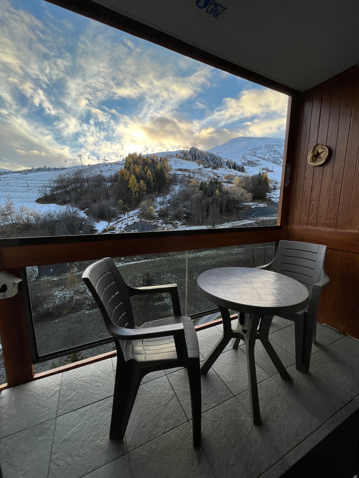 A balcony features a round table and two chairs with a scenic view of a snow-covered landscape. Tall trees are visible in the distance, framed by a sky with soft clouds reflecting warm hues.