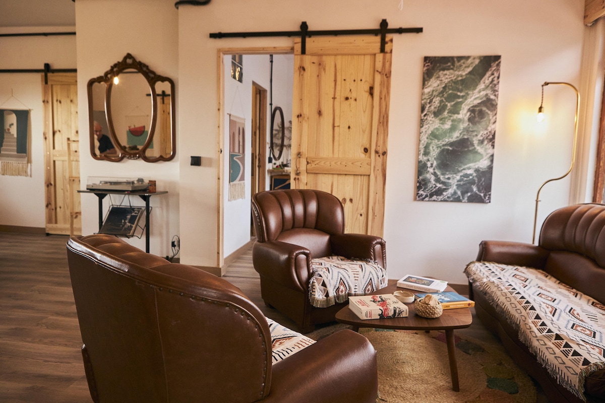 A cozy living area features two brown leather armchairs and a matching sofa adorned with patterned throw pillows. A round coffee table holds a few books and magazines. Light wood sliding doors lead to other spaces, with artwork displayed on the walls, enhancing the inviting ambiance.
