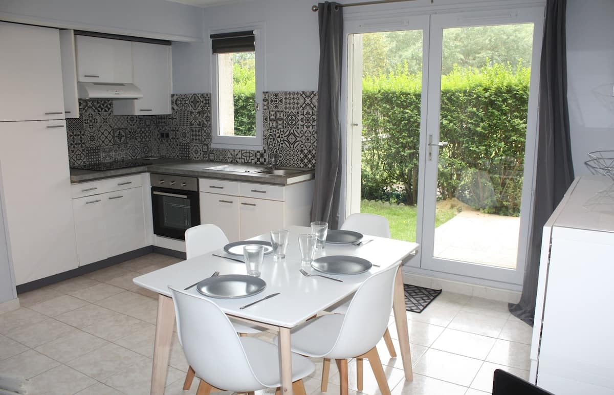A modern kitchen and dining area is equipped with sleek cabinets and countertops. A table set for four displays dinnerware, complemented by white chairs. Large windows allow natural light to illuminate the space, revealing views of the green garden outside.