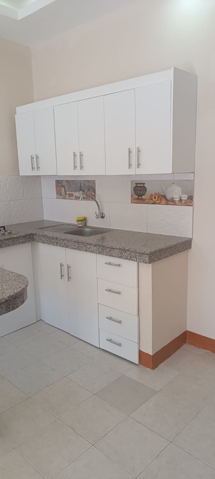 A clean kitchen features white cabinetry paired with a light granite countertop. The sink is positioned beneath a tile backsplash, and kitchen essentials are visible on the counter. A circular surface is shown with a simple design, contributing to a functional layout.