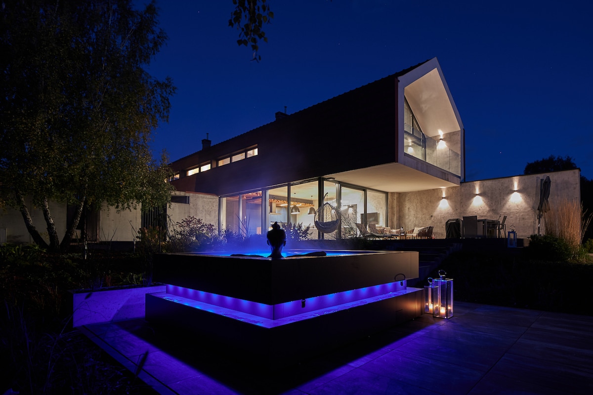 A modern home is illuminated at night, featuring expansive windows that reveal a stylish interior. An outdoor jacuzzi is highlighted with soft blue lighting, accompanied by decorative lanterns. The surrounding area is lush with greenery, enhancing the peaceful ambiance.