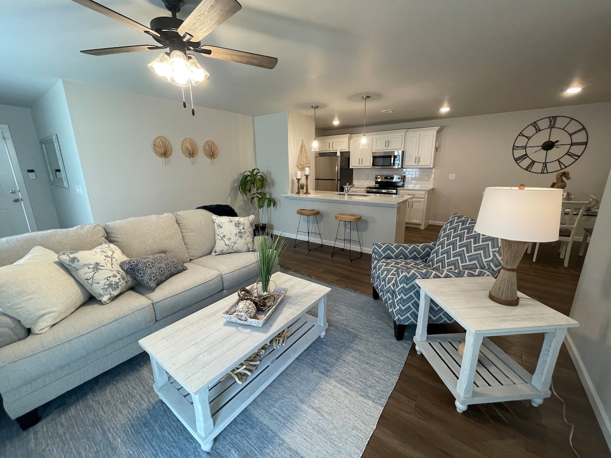 A bright living area features a light-colored sofa adorned with patterned cushions. A white coffee table with decorative elements sits centrally. To the side, a cozy chair with chevron fabric complements the space, with a floor lamp providing soft illumination. An open kitchen area is visible in the background.