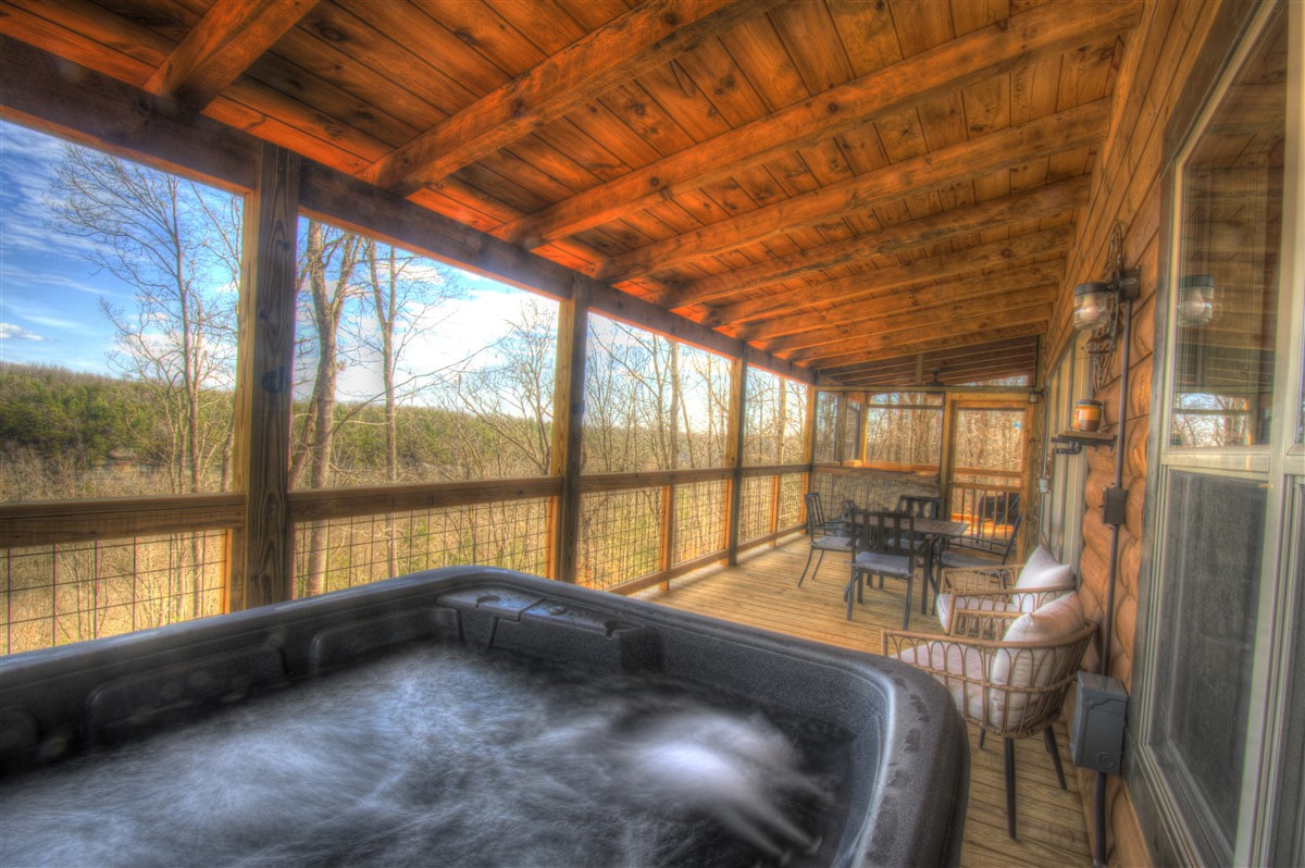 The covered deck features a hot tub with bubbling water, surrounded by wooden railings and large windows that provide expansive views of the surrounding woodlands. A dining table with chairs and a cozy armchair are also present, creating a comfortable outdoor space.