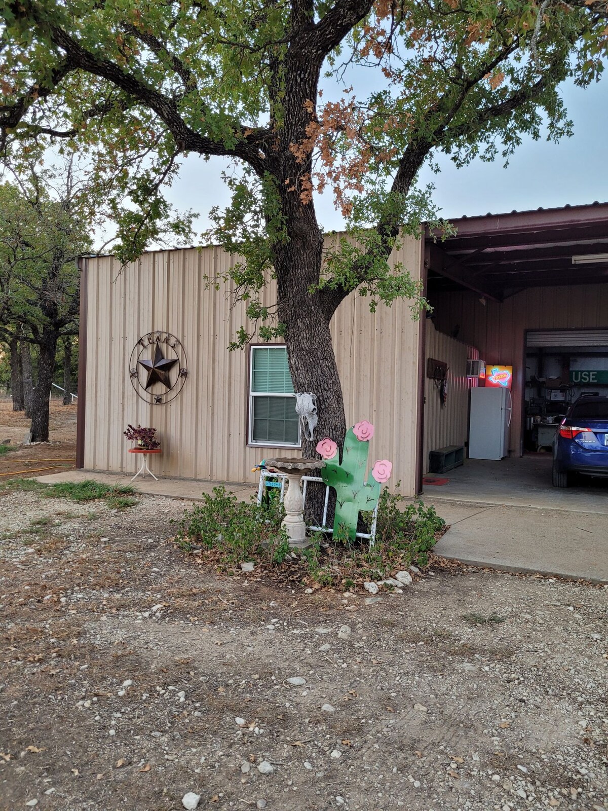 The exterior of the barndominium-style guest house features a metallic façade and a decorative star. Surrounding greenery includes a cactus with pink flowers and a small garden area. A porch area is visible, and a vehicle is parked nearby.