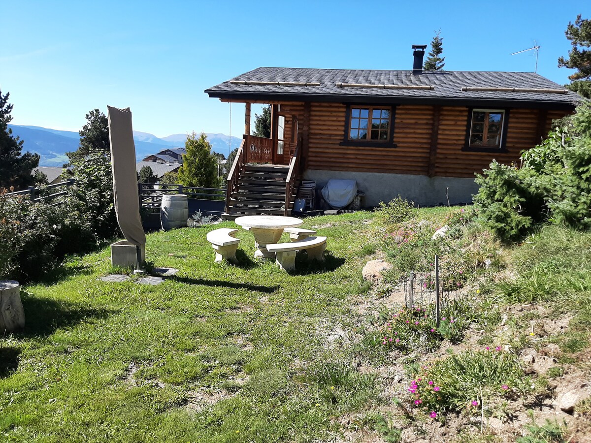 A wooden chalet is situated on a grassy area with a stone seating arrangement in front. Surrounding the chalet are shrubs and trees, while the landscape features distant mountains under a clear blue sky.