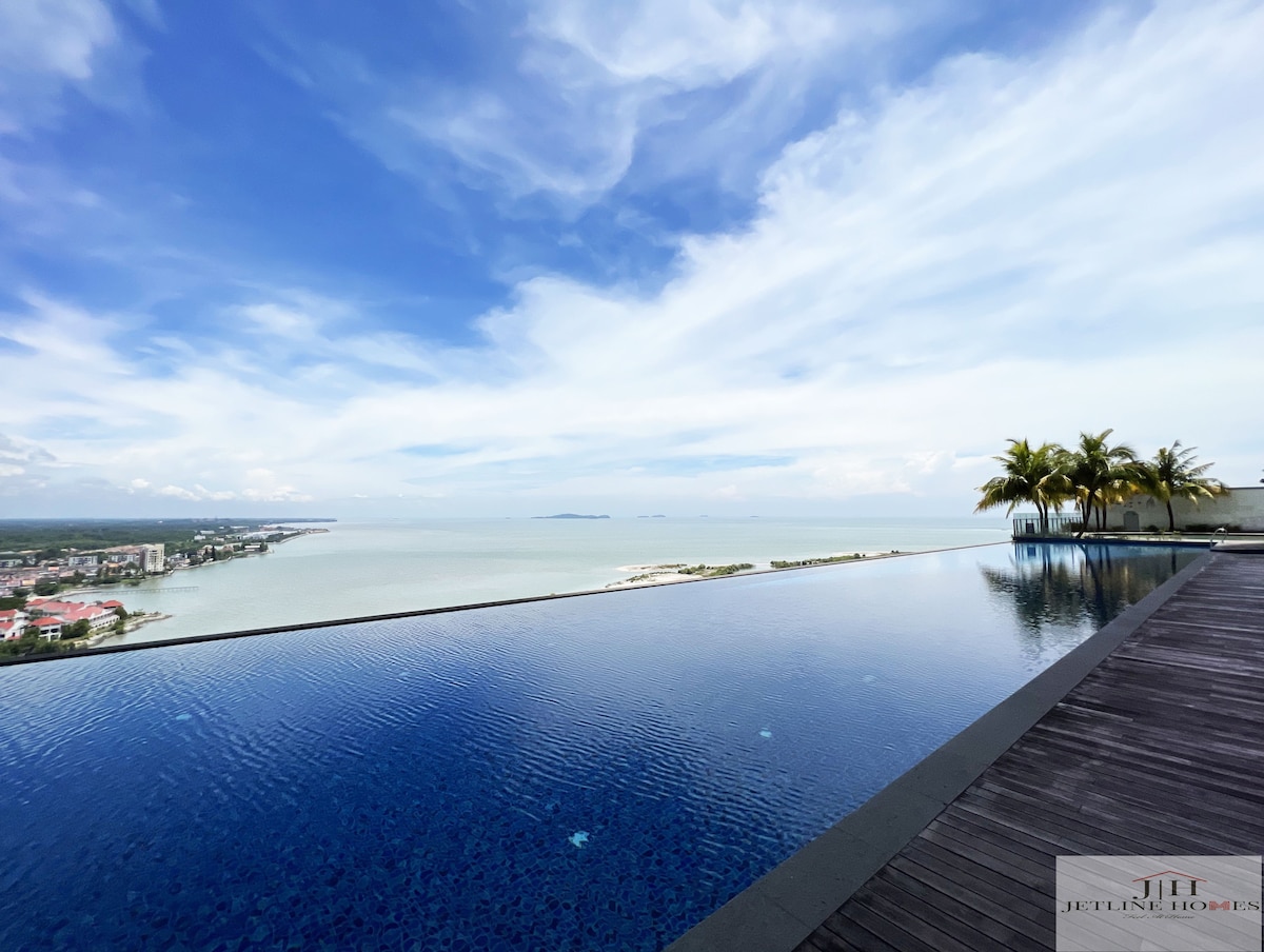 An infinity pool extends towards the horizon, reflecting the sky and surrounding sea. Palm trees frame the edge, while the tranquil water contrasts against the cloud-dotted sky, creating a serene atmosphere.