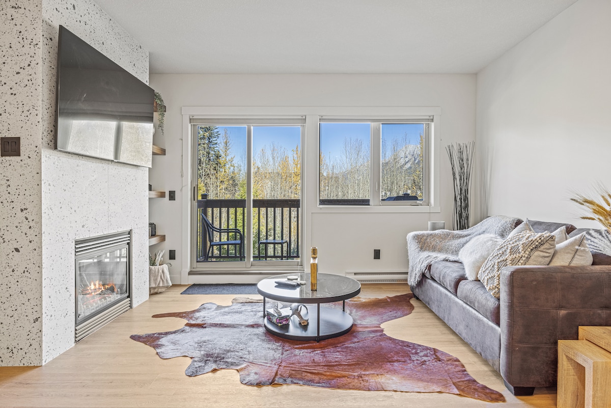 A spacious living area features a cozy sofa positioned near a contemporary fireplace. Large windows offer views of the surrounding mountains and outdoor balcony. A round coffee table rests on a decorative area rug, enhancing the inviting atmosphere of this modern retreat.