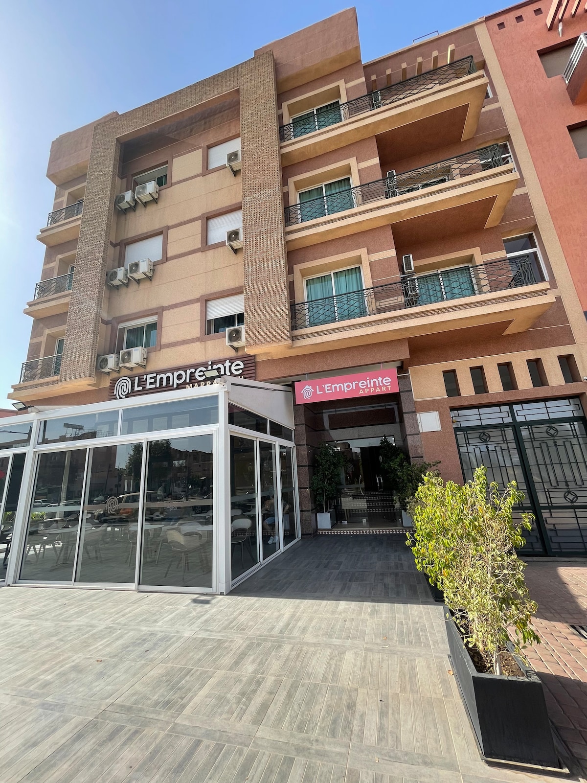 The exterior of L'Empreinte appears inviting with large glass windows and a modern facade. Balconies are visible on the upper floors, while a shaded entrance area features greenery and seating options. The surroundings include a paved walkway, enhancing access to the establishment.