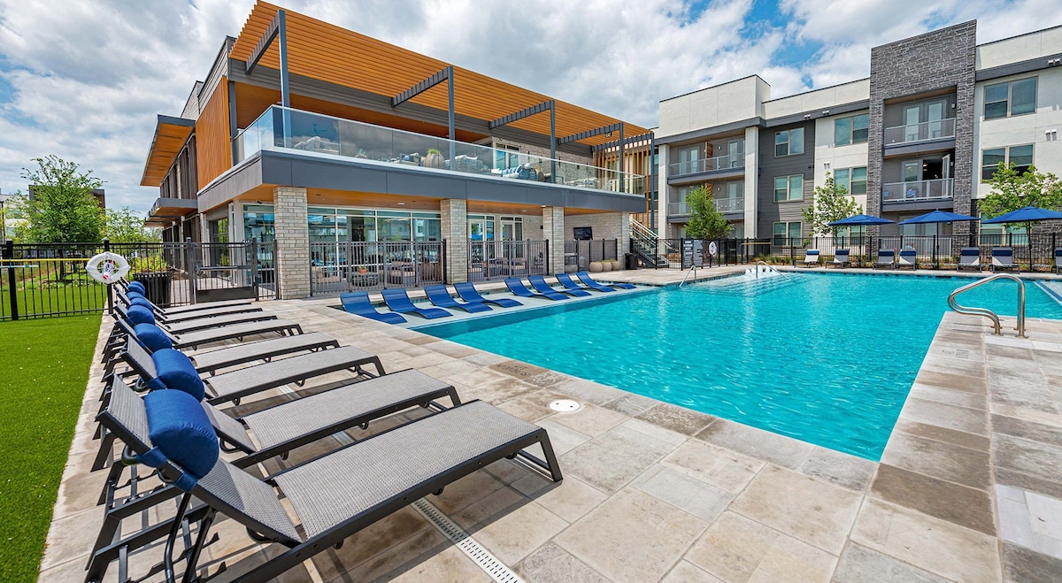 The image captures a resort-style pool surrounded by lounge chairs in a spacious area. A modern clubhouse with shaded outdoor seating overlooks the pool. Lush green landscaping and clear skies enhance the ambiance, creating a relaxing environment for guests.