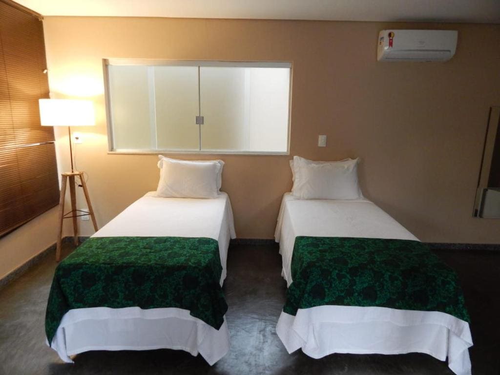 Two spacious twin beds are positioned side by side, each adorned with green bed covers. A minimalist floor lamp stands beside one bed, and natural light enters through a nearby window, providing a bright and airy environment. A wall-mounted air conditioning unit is visible above.