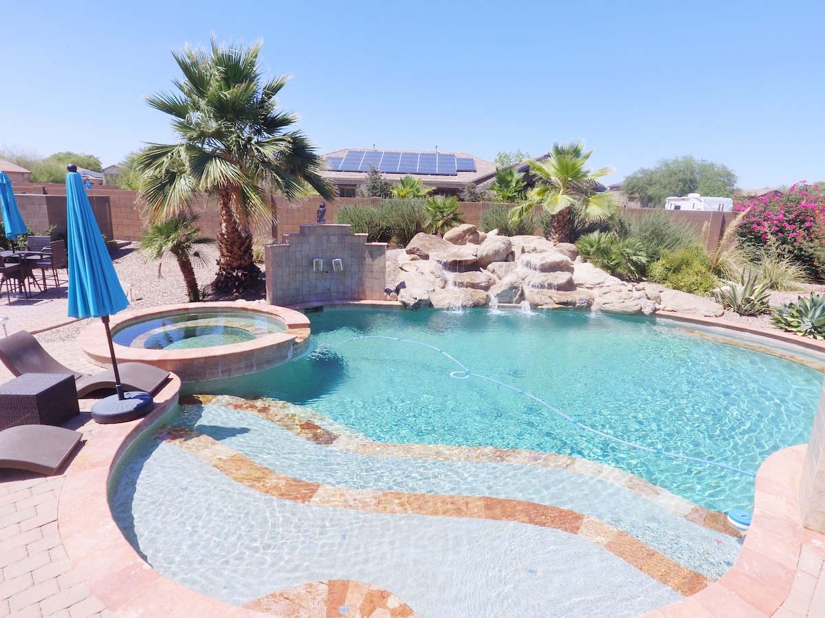 A sparkling pool is framed by natural stone and tropical palm trees, creating a serene outdoor space. Blue umbrellas provide shade for stylish lounge chairs. The area is beautifully landscaped with desert flora, offering a private oasis for relaxation.