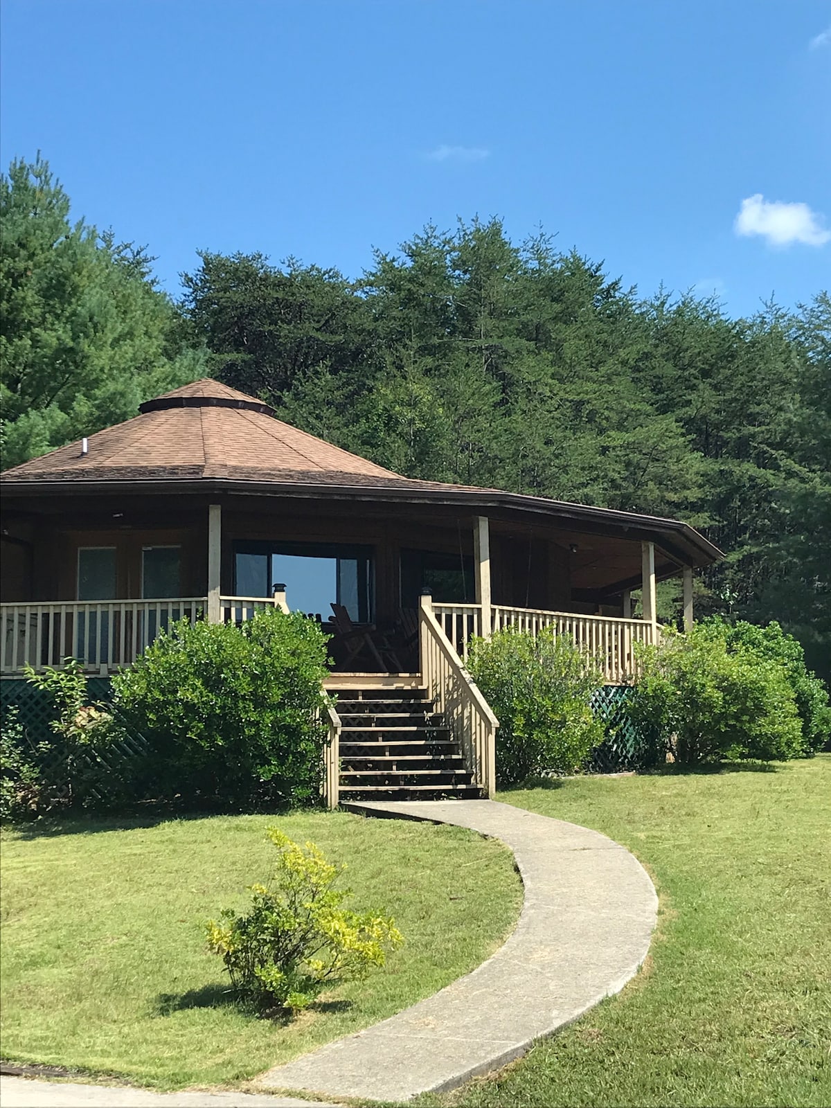 The Roundhouse features a circular design with a sloping roof, surrounded by lush greenery. A pathway leads to the wooden porch with steps that invite guests up to the entrance. Large windows allow natural light to brighten the interior.