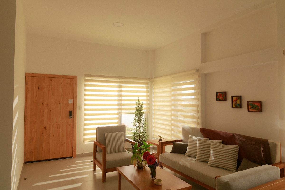 A light-filled living area features two comfortable chairs and a plush sofa, complemented by a coffee table that hosts a floral arrangement. Soft shades cover large windows, allowing natural light to filter in, while a wooden door provides entry to the space.