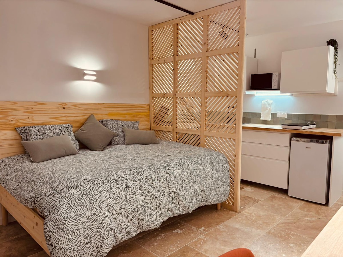 A spacious sleeping area features a large bed topped with a patterned duvet and several cushions. A wooden partition divides the space from a modern kitchenette equipped with essential appliances, including a refrigerator and a microwave, complemented by tiled backsplash.