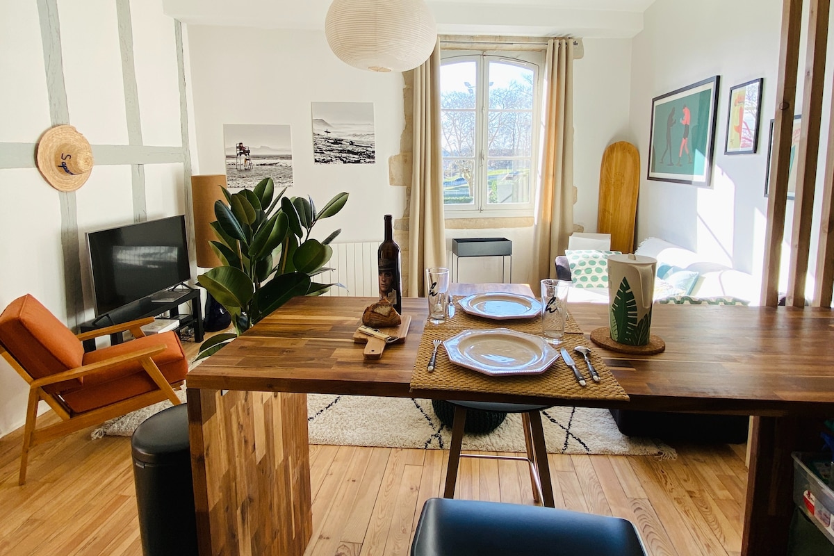 A well-appointed living area is displayed with a wooden dining table set for two, featuring plates and utensils. A cozy armchair, various artworks, and a plant enhance the inviting space, while a large window allows natural light to illuminate the warm wood flooring.