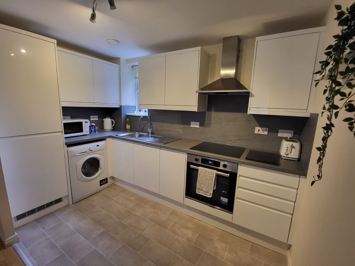 A modern kitchen is equipped with sleek white cabinetry, gray countertops, and stainless steel appliances. A washing machine is positioned beside the sink, and a microwave sits on the counter. Soft natural light brightens the space through a nearby window.
