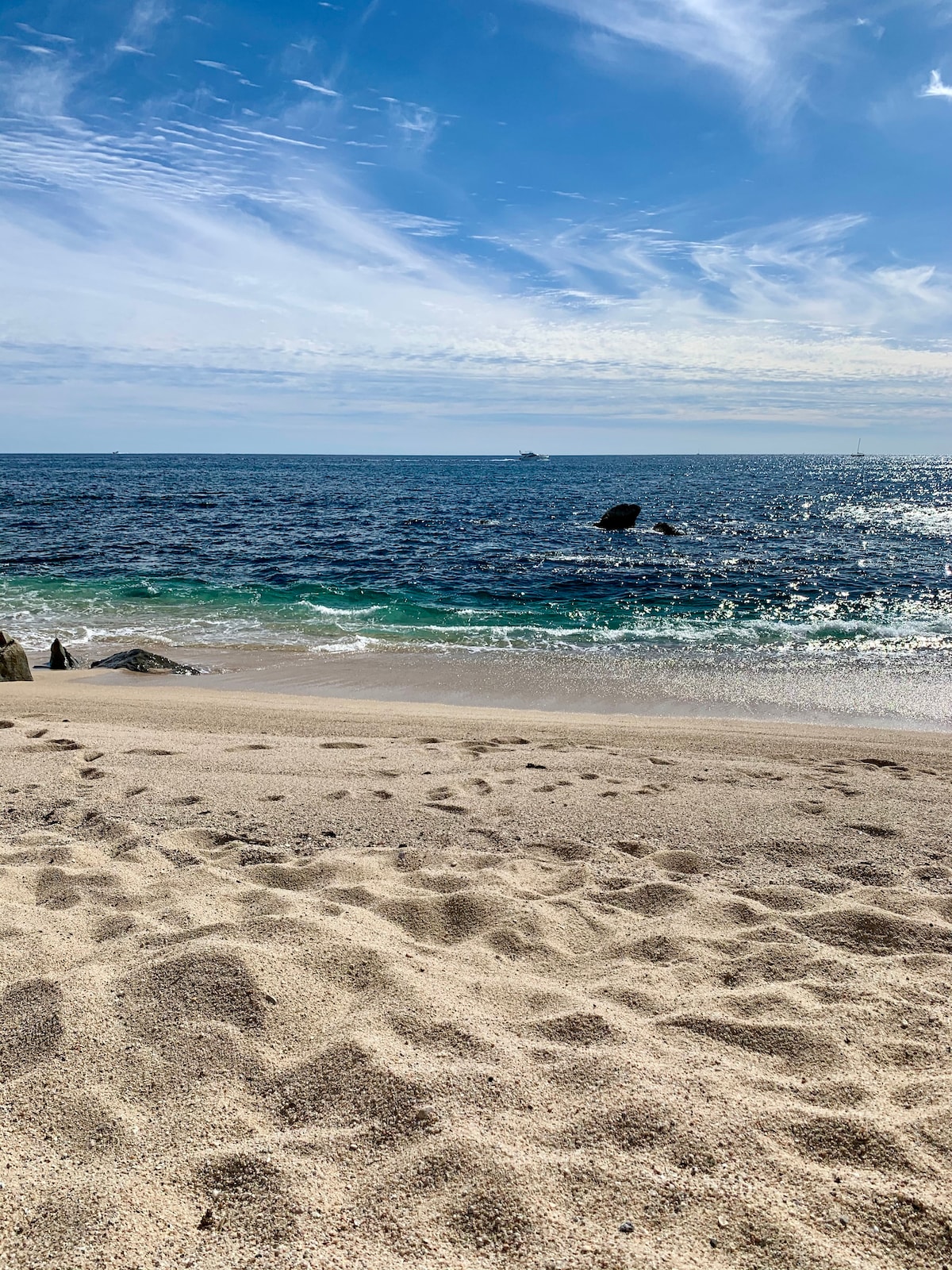 A scenic beach view captures gentle waves lapping against a sandy shore. The horizon showcases a blend of blue hues from the ocean sky and calm waters. A distant rock formation is visible just offshore, adding depth to the peaceful waterfront landscape.