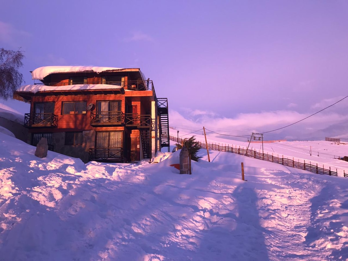 A two-story wooden building is set against a backdrop of snow-covered terrain. The structure displays multiple balconies and large windows, reflecting the soft hues of the sunset. The pathway leading to the entrance is surrounded by deep snow, enhancing the winter atmosphere.