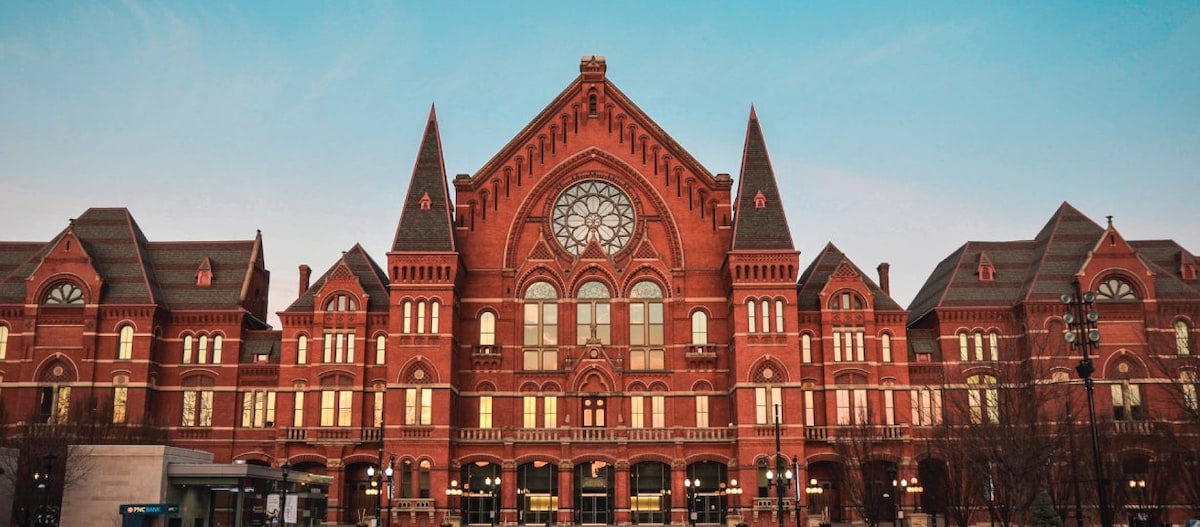 The historic building showcases intricate Gothic architecture with prominent red brick, steeply pitched roofs, and ornate window designs. A large round window dominates the center, framed by towers on either side. The soft glow of lights reflects on the facade, highlighting the building's detailed craftsmanship.