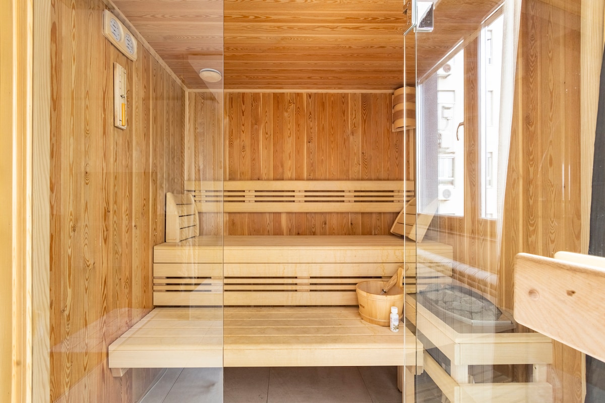 A modern sauna space is enclosed by clear glass panels. The interior features warm wooden walls and seating, with a wooden bucket and ladle positioned on the floor. Bright light filters in through a window, enhancing the inviting atmosphere.