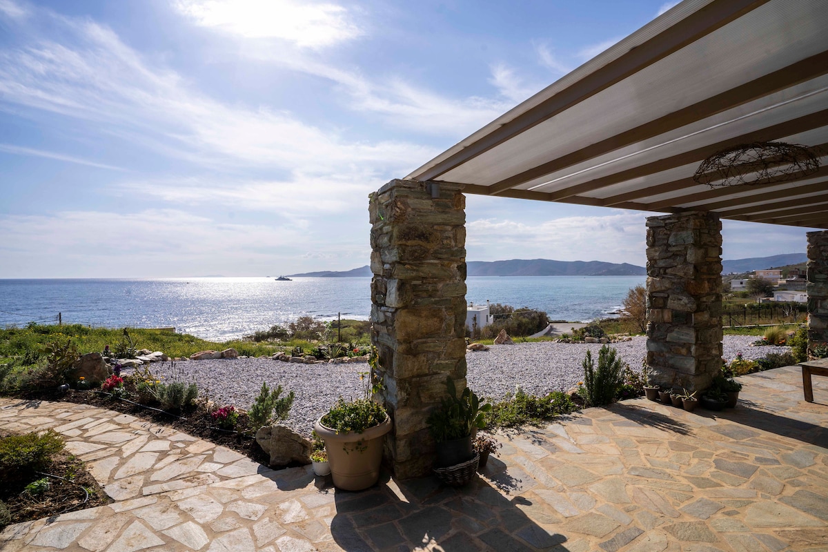 The exterior space features a stone-covered patio with a view of the serene sea. Colorful plants are arranged in pots, while the sky above showcases soft clouds and sunlight reflecting off the water. A shaded area is created by a roof structure, enhancing outdoor relaxation.