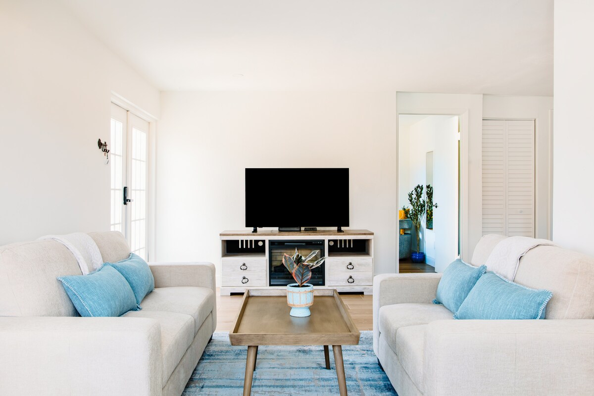 A light-filled living area features two neutral sofas adorned with blue accent pillows, positioned across from a sleek entertainment console with a television. A wooden coffee table sits in the center, complemented by a decorative plant. Glass doors provide access to the outside.