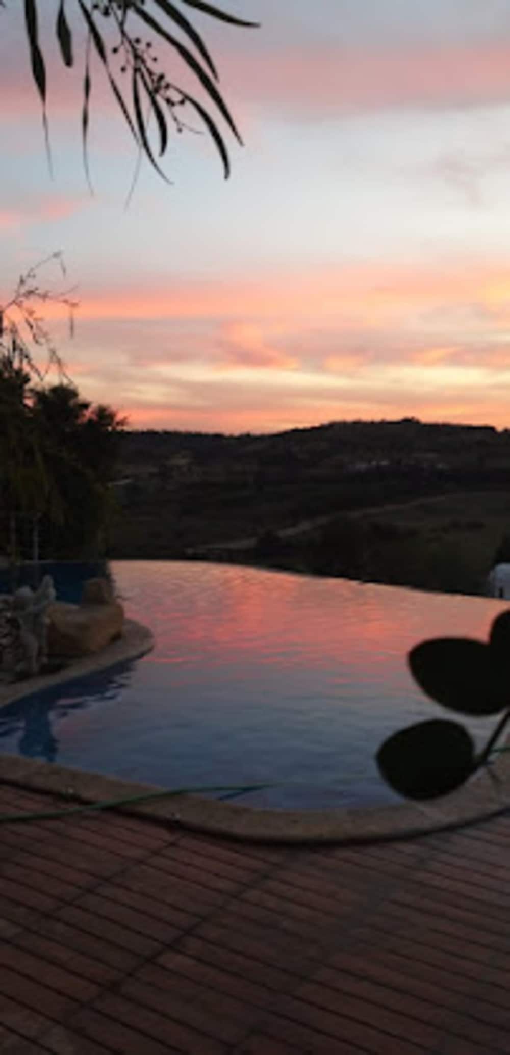 An infinity pool reflects the pastel colors of a sunset, surrounded by gentle hills and greenery. The tranquil scene is framed by leaves, creating a peaceful ambiance for relaxation.