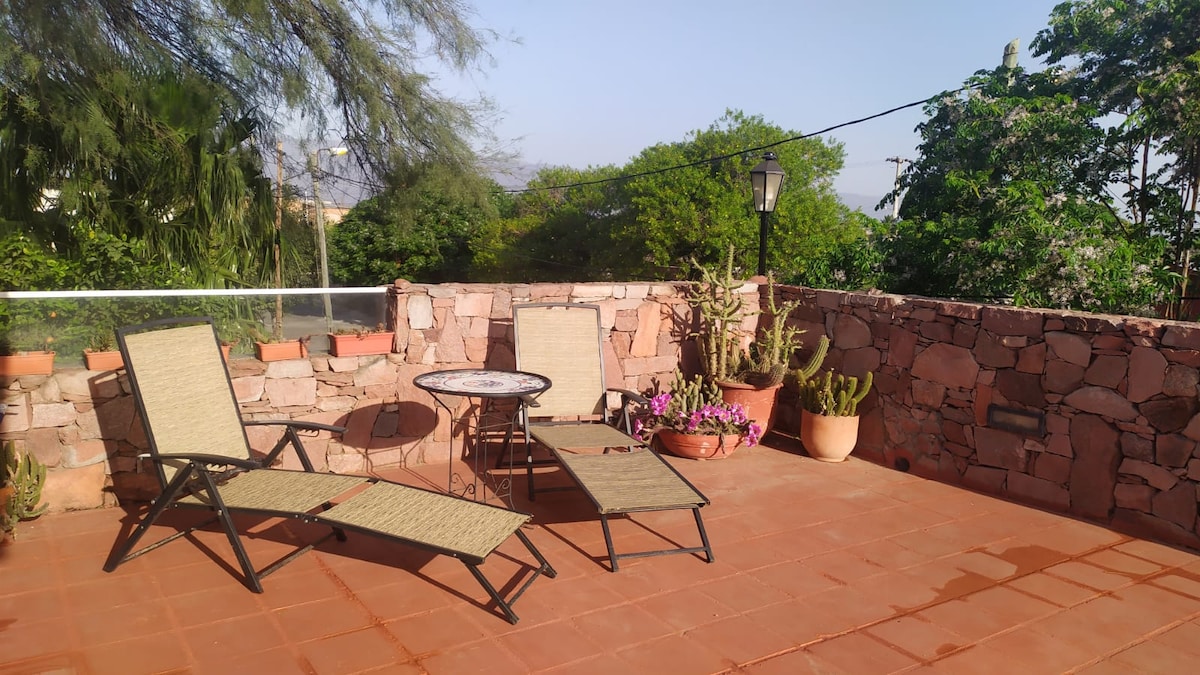 A serene terrace offers two reclining chairs and a small round table surrounded by lush greenery and flowering plants. The natural stone wall provides privacy and warmth, while a gentle light illuminates the space. This outdoor area is ideal for relaxation and enjoying fresh air.