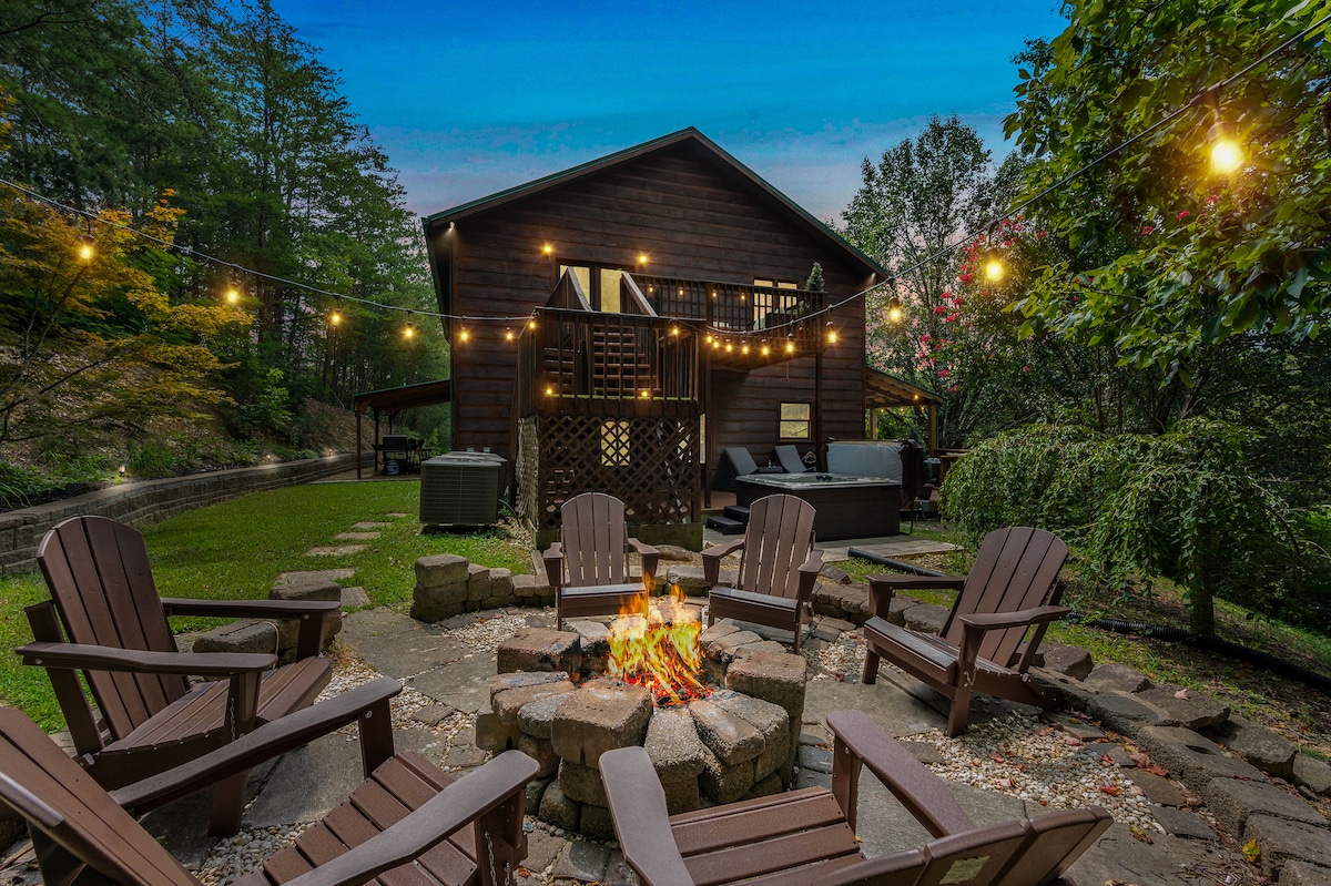 An outdoor fire pit area features Adirondack chairs arranged in a circular formation around a stone fire pit. String lights are hung above, creating an inviting atmosphere. The cottage's exterior is visible in the background, surrounded by trees and lush greenery.