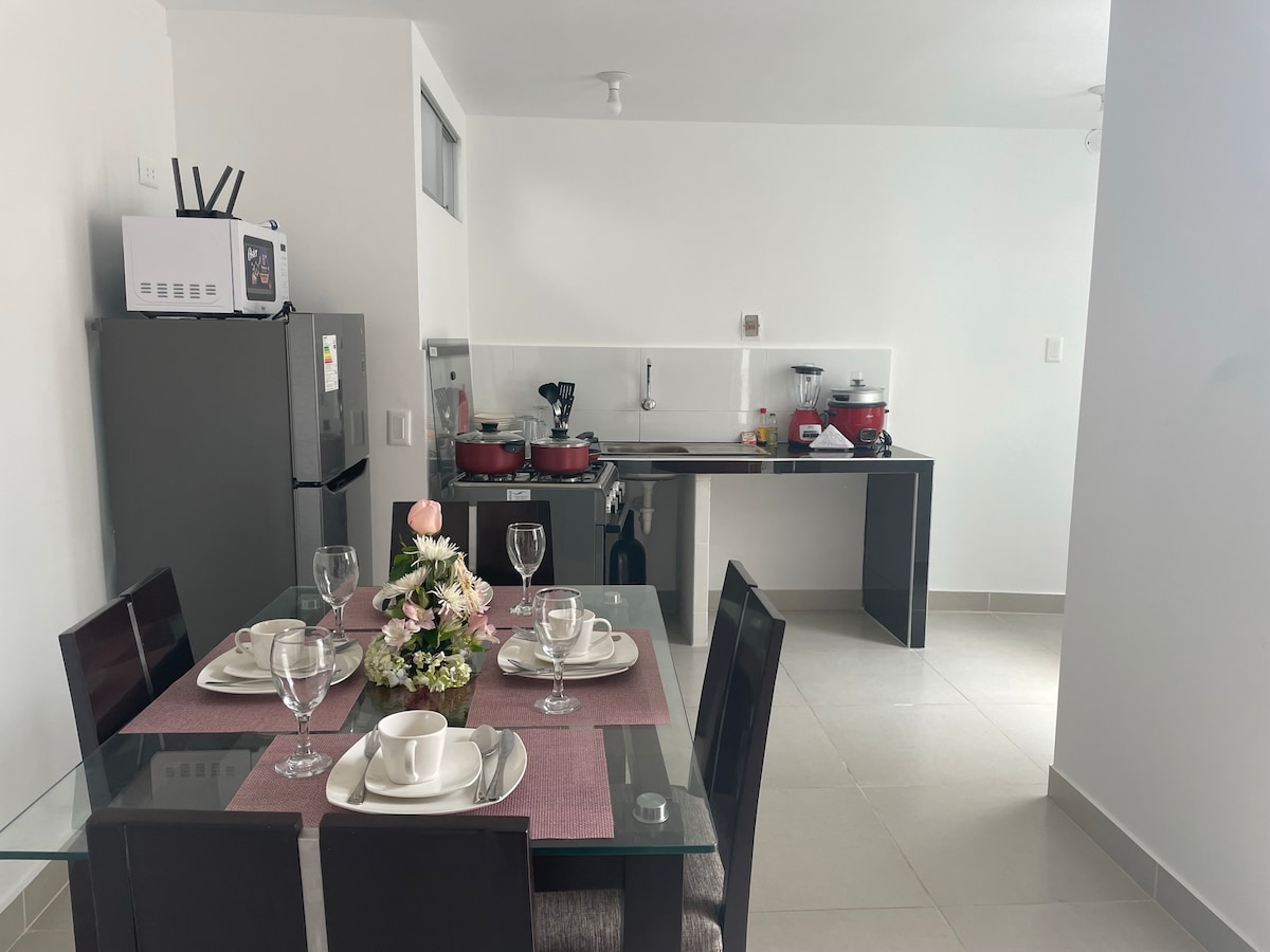 A modern kitchen and dining area is shown, featuring a glass dining table set for four. A refrigerator and cooking appliances are visible, with pots displayed on the countertop. The space is illuminated by bright, neutral lighting, enhancing the clean, minimalist design.