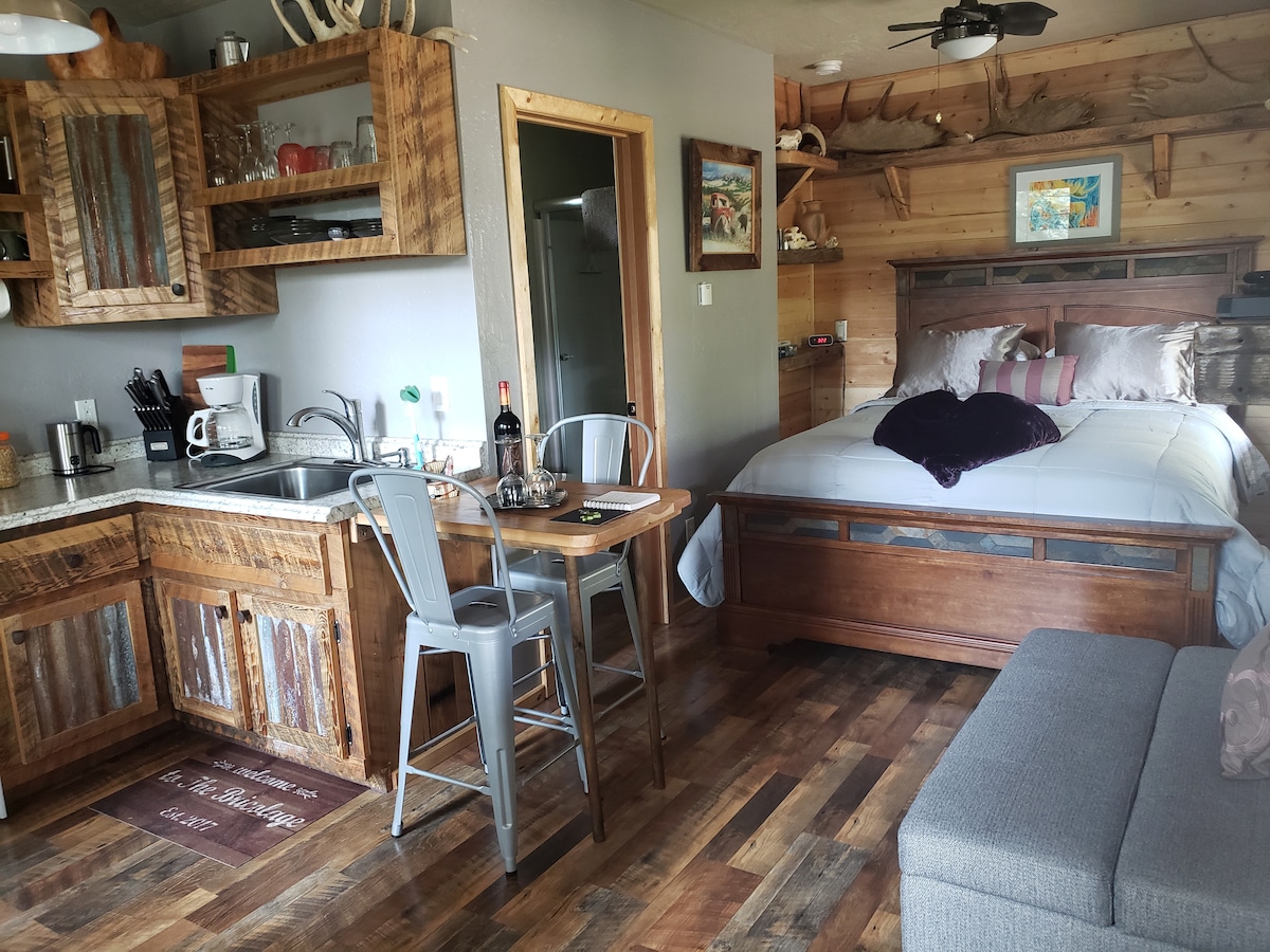 A cozy cabin interior features a queen-sized bed with a rustic wooden frame and a small dining table with metal chairs. The kitchen area includes essential appliances and wooden cabinetry, while a comfortable sofa is positioned nearby. Natural light fills the space through a nearby window.