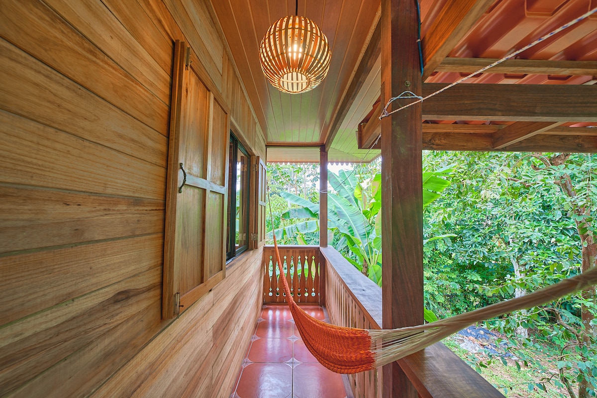 A wooden balcony is seen with warm tones, featuring a vibrant orange hammock hanging between support beams. Windows with wooden shutters allows a view of the lush greenery. A woven pendant light hangs from the ceiling, adding a cozy touch to the outdoor space.