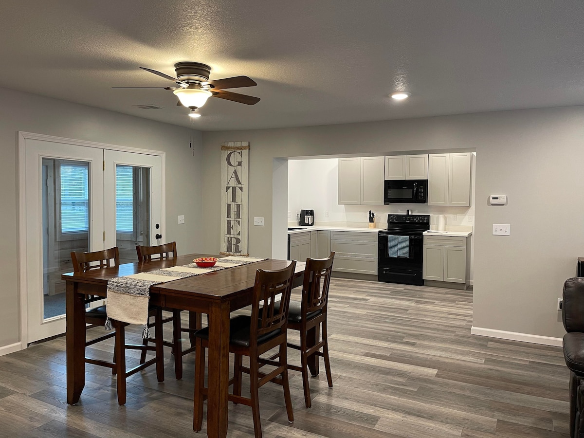 An open dining area features a rectangular wooden table surrounded by high-backed chairs, illuminated by overhead lighting. A decorative sign reading 'GATHER' is displayed on the wall. The adjacent kitchen is visible, equipped with modern appliances and light cabinetry.
