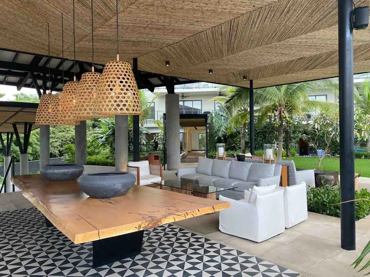 A spacious outdoor living area features a large wooden table with a dark base, surrounded by white cushioned seating. Unique woven light fixtures hang above, enhancing the atmosphere. Lush greenery and palm trees create a tropical backdrop, adding to the inviting environment.