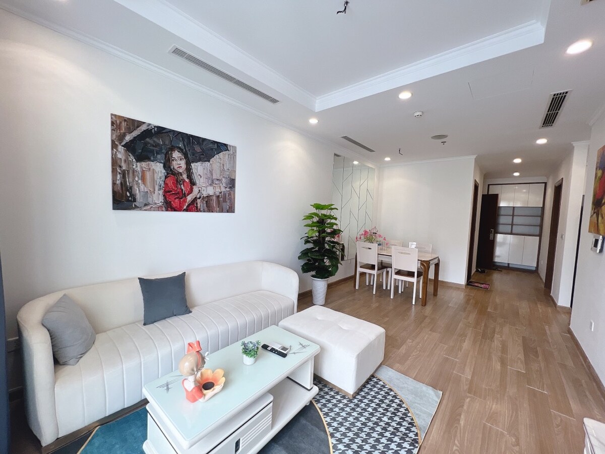 A light-filled living area features a white sofa alongside a stylish coffee table. A small plant adds greenery, while a dining table with chairs occupies the adjacent space. Artwork adorns the wall, contributing to the room's aesthetic appeal, and a doorway leads to further areas.