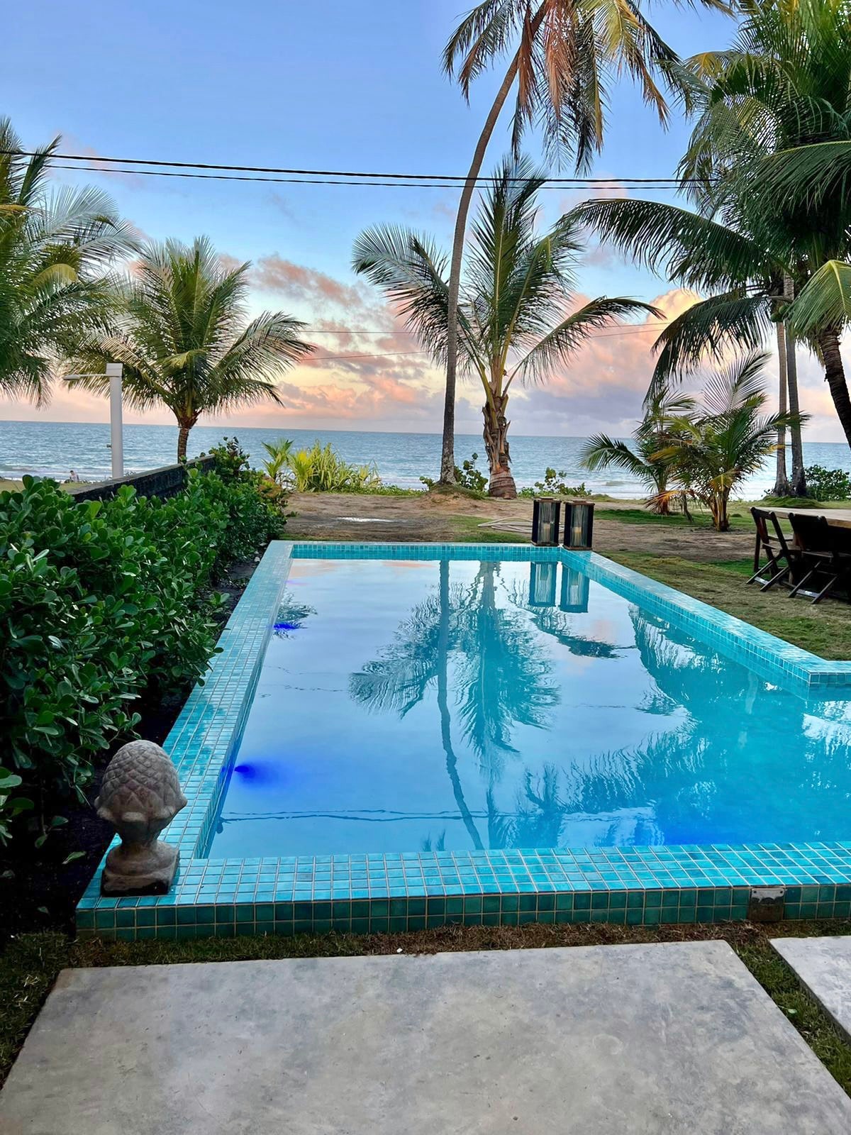 A serene outdoor space features a clear swimming pool surrounded by lush greenery and palm trees. The tranquil water reflects the sky's soft pastel colors, showcasing a peaceful coastal view in the background.