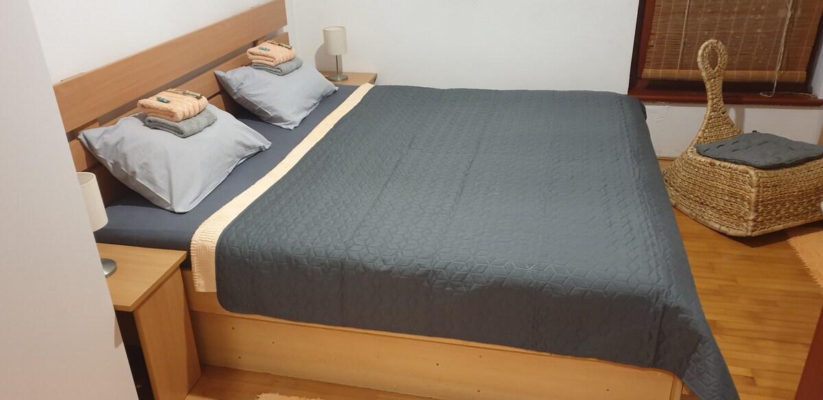 A spacious bed covered with a gray quilted blanket is presented, adorned with two neatly arranged pillows. A light-colored bed frame and nightstands are featured on either side, with minimalistic lamps placed atop each. A wicker chair and window with natural light complete the room.