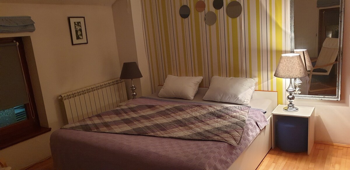 The sleeping area features a spacious bed adorned with a patterned blanket. Two bedside lamps sit on each side, complemented by a large mirror and a colorful striped accent wall. Natural light filters through a window with light-colored curtains.