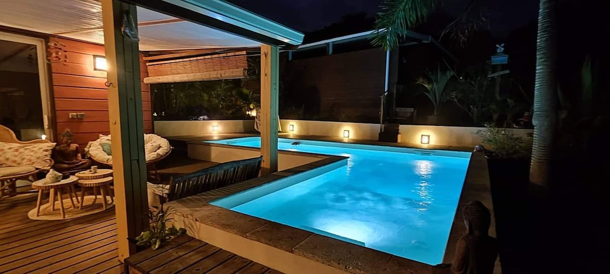 The outdoor area features a well-lit swimming pool surrounded by wooden decking. Comfortable seating arrangements and ambient lighting create a serene environment. Lush greenery and decorative elements enhance the tranquil setting, inviting relaxation under the night sky.