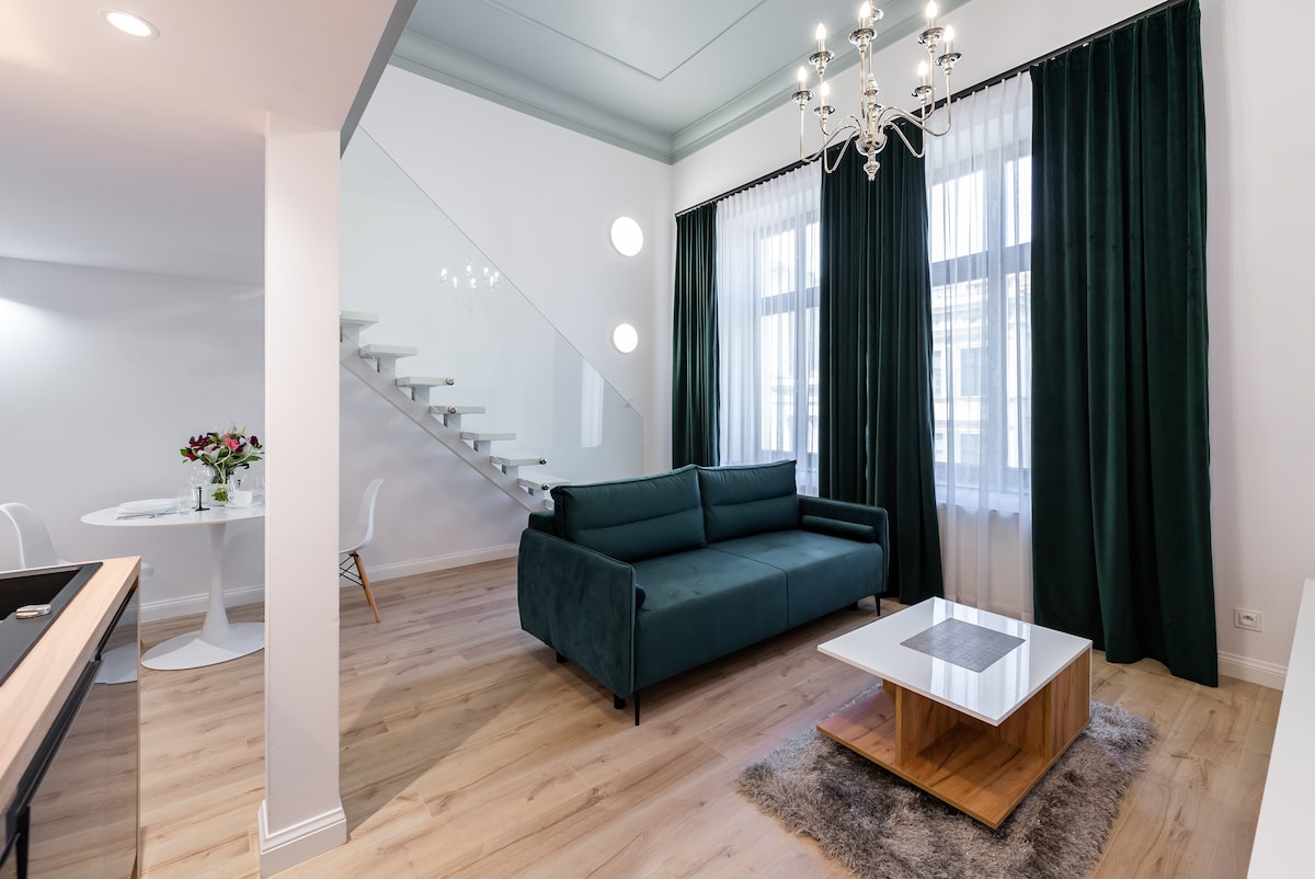 A modern living area features a plush green sofa positioned near large windows with sheer curtains. A sleek wooden coffee table sits on a light rug, while a circular dining table with white chairs is visible in the background, enhancing the open layout.