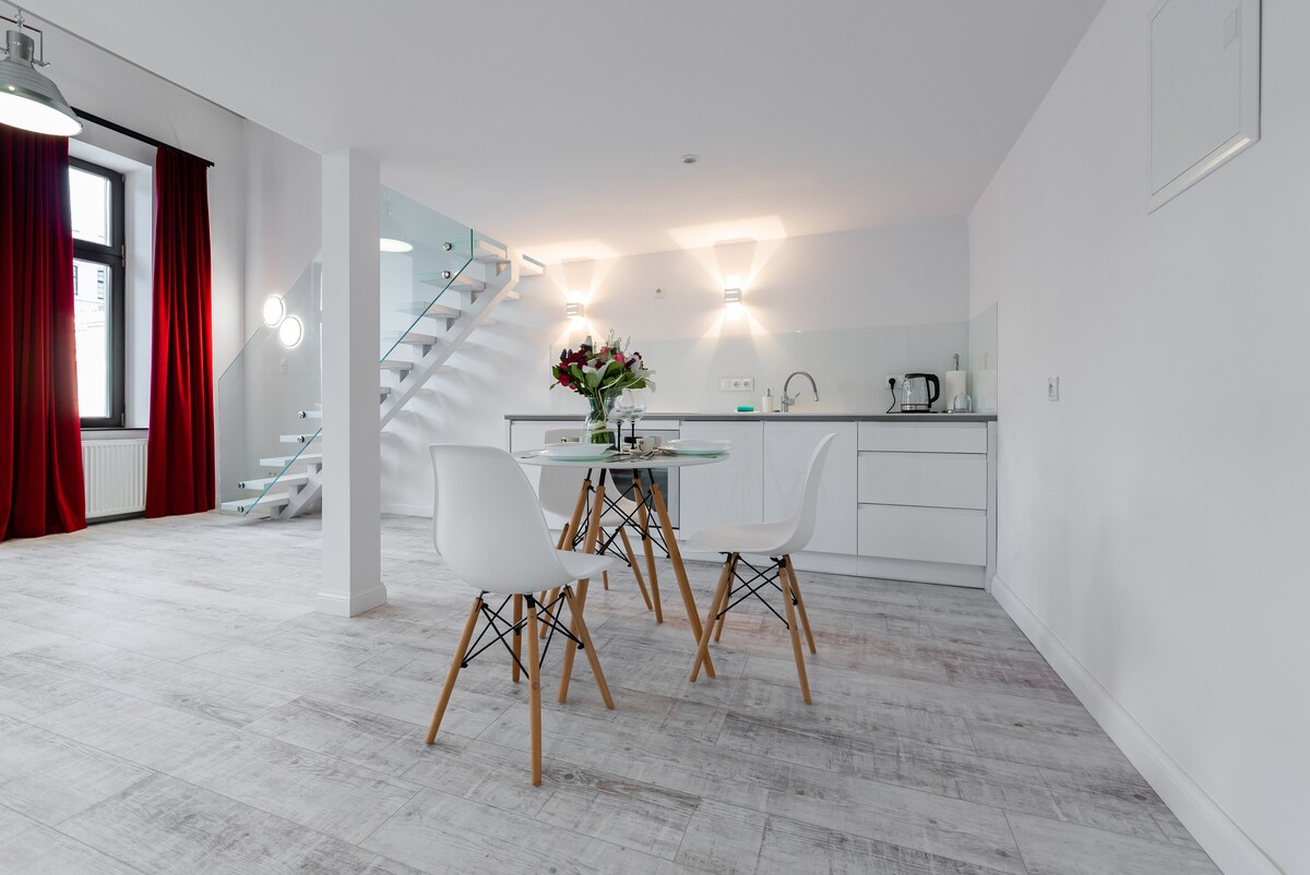 An open dining area features a glass table surrounded by three white chairs. The modern kitchen is visible in the background, equipped with essential appliances. Tall windows with red curtains allow natural light to fill the space, while a staircase leads to the upper level.