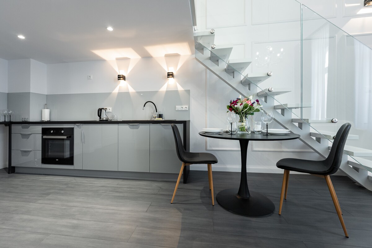 A modern kitchen area is visible, featuring sleek grey cabinets and a built-in oven. A round dining table with two black chairs is situated nearby, complemented by a vase of fresh flowers. A glass staircase adds an elegant touch to the space.