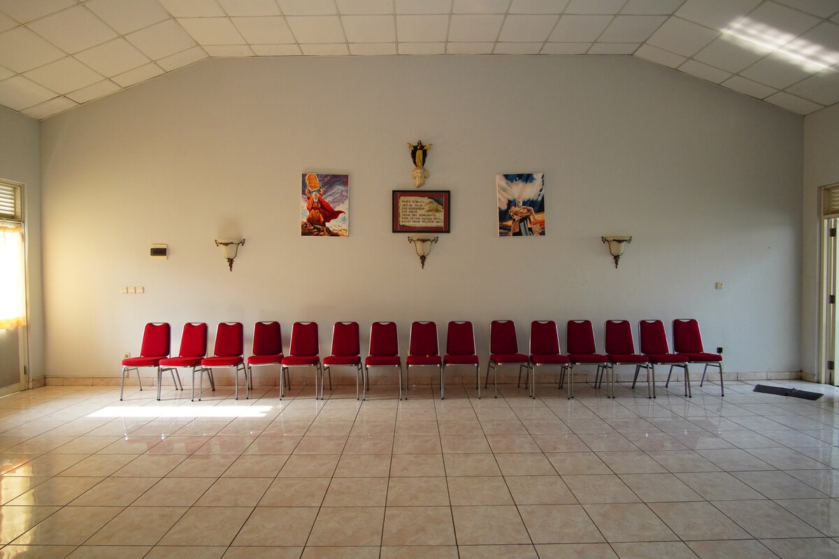 A spacious hall is showcased with a row of red chairs neatly arranged along one wall. The glossy tiled floor reflects the soft natural light coming from adjacent windows. Art pieces are displayed on the wall above, contributing to the room's purpose.